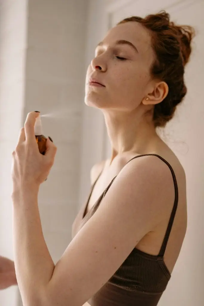 Person holding a brown glass spray bottle, misting product onto their neck and upper chest, showcasing a skincare or fragrance application in a soft, natural lighting setting.
